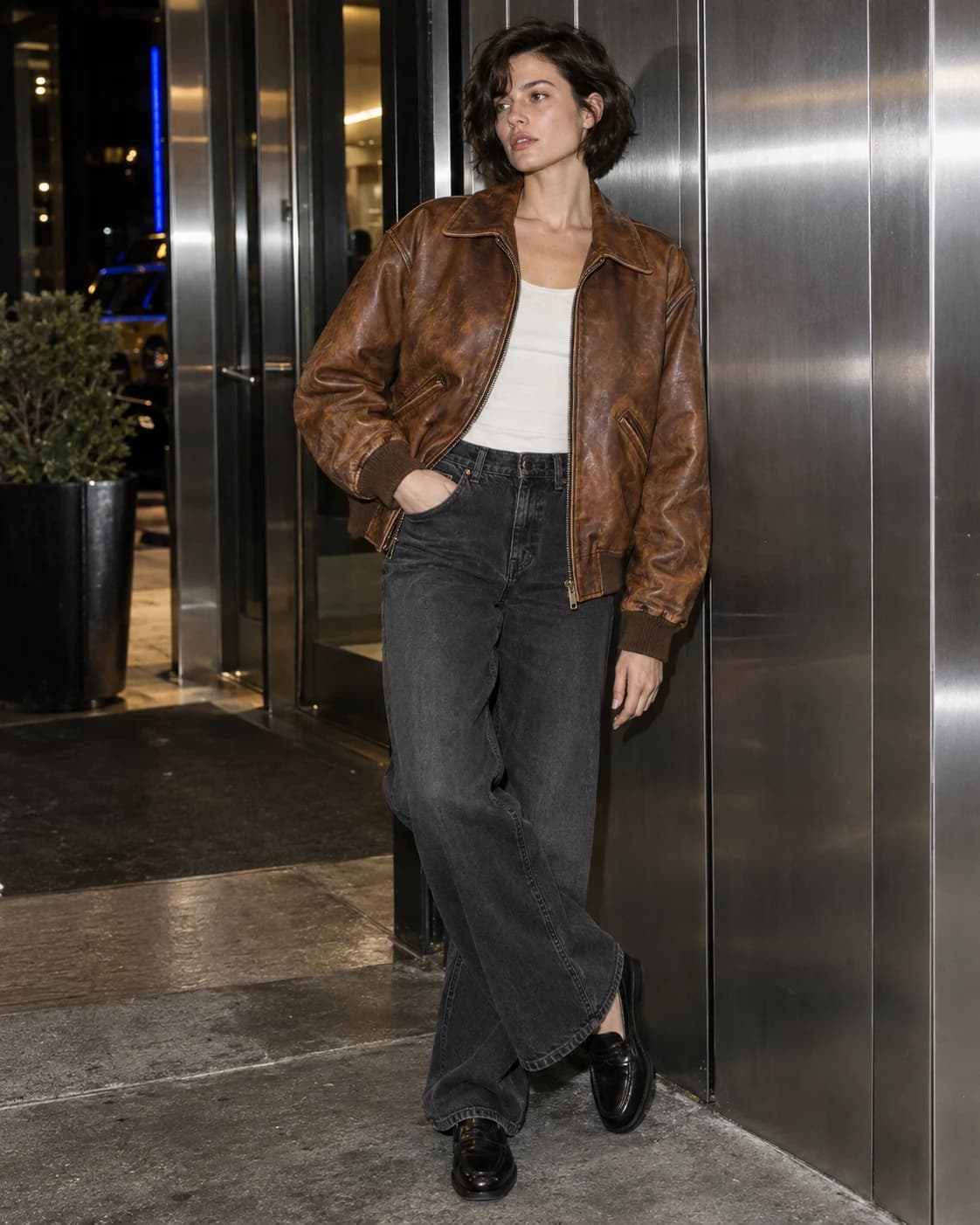 Model im cognacbraunen Lederblouson mit weiter dunkler Jeans vor einer Metallfassade — KI-generiertes Beispiel